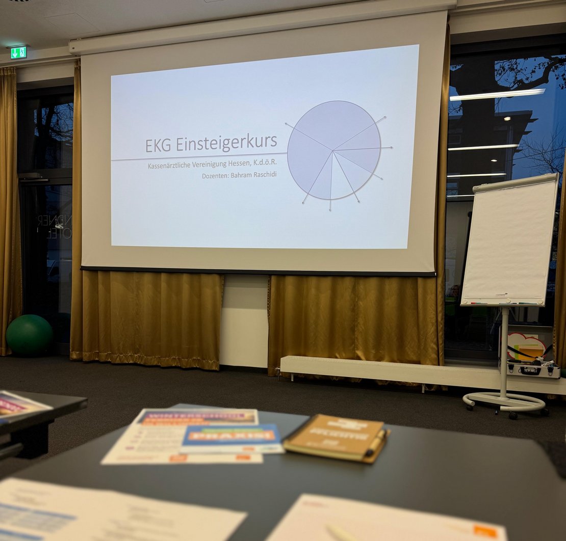Auf einer Präsentation ist der Titel EKG-Kurs zu lesen. Vorne dran steht ein Tisch mit Stiften und Blöcken.