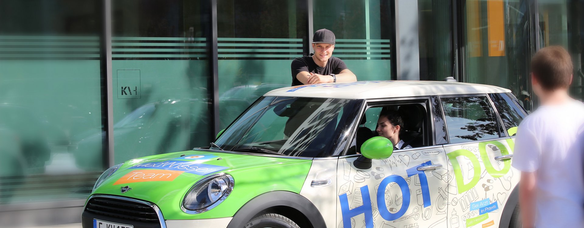 Mini Cooper im Kampagnendesign Sei Ärztin Praxis Leb Hessen. Frau sitzt im Auto. Man lehnt sich auf das Auto